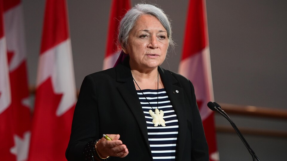 Mme Simon parle dans un micro installé devant des drapeaux du Canada.