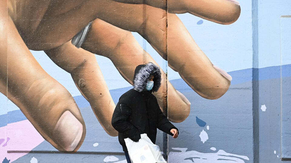 Un passant masqué marchant devant une main gigantesque sur une fresque murale.