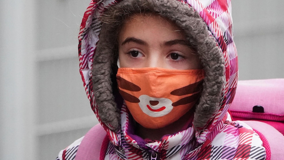 Une fillette arrive à l'école, un masque orange sur le visage et le capuchon de son manteau d'hiver sur la tête.