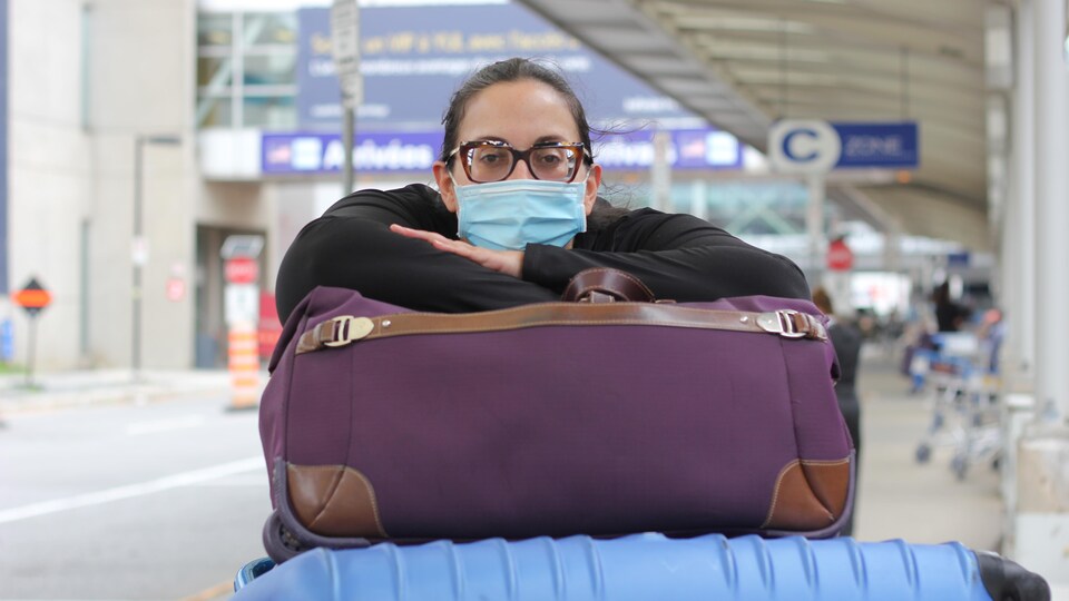 Une femme les bras croisés sur une pile de valises à l'aéroport. Elle porte des lunettes et un masque bleu poudre sur le nez et la bouche. 