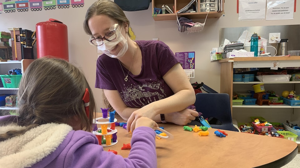 Une femme porte un masque à fenêtre et sourit. Elle est assise à une table avec une enfant et elles manipulent de petites figurines.