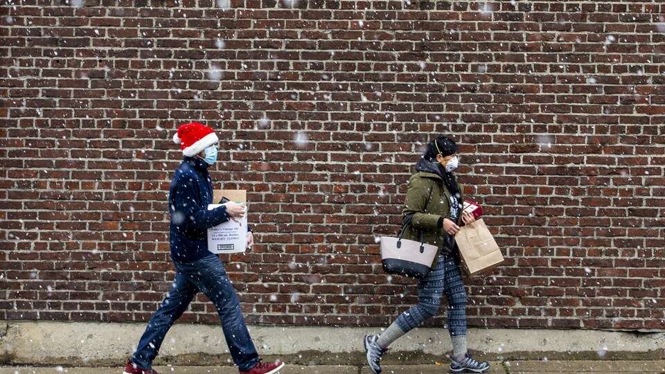 Deux personnes marchent sous la neige avec des sacs de magasinage à Toronto le 22 novembre 2020. L'une d'elles est coiffée d'une tuque de Noël.