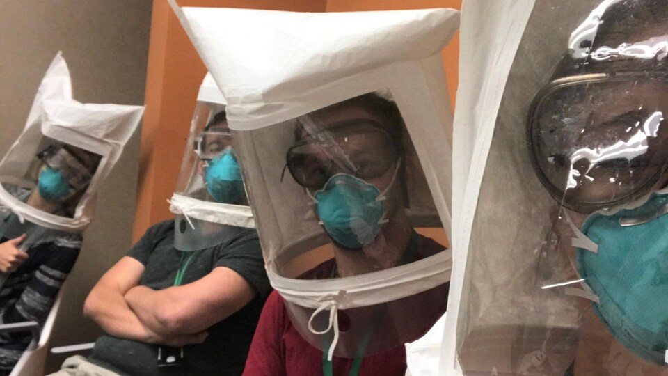Quatre hommes sont assis sur des chaises dans un hôpital avec d'énormes masques blancs qui protège leur tête au complet.