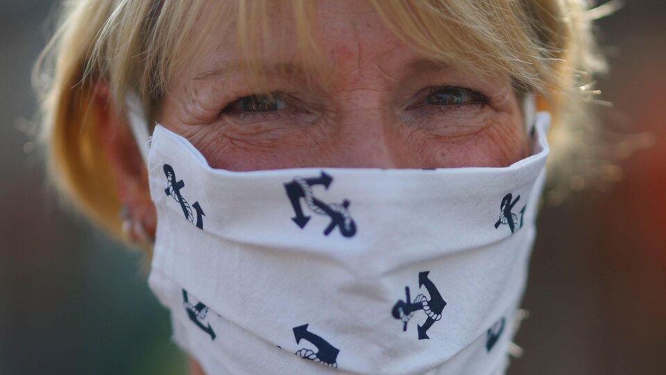 Une femme aux cheveux blonds courts a sur son nez et sa bouche un masque en tissu blanc avec des motifs d'ancres de bateau. 