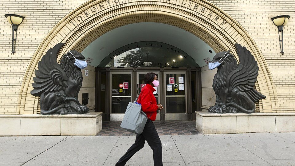 Une femme portant un masque passe devant une bibliothèque où deux statues d'aigles ont un aussi un couvre-visage.