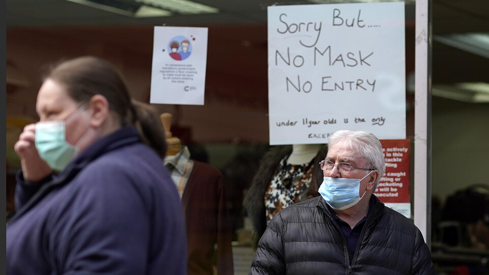 Un homme et une femme portant des masques de protection  avec en arrière-plan une pancarte avisant les clients qu’ils ne seront pas servis s’ils ne portent pas de masque.