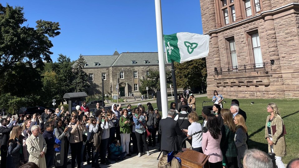 Des dizaines de personnes et dignitaires sont rassemblées devant un mât avec un drapeau franco-ontarien en train d'être levé.