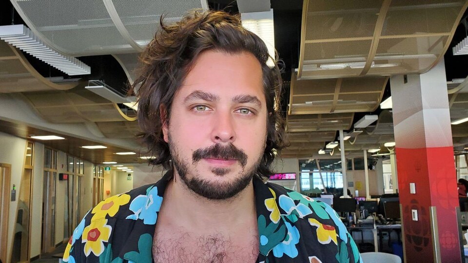 Un homme aux cheveux longs et chemise fleurie déboutonnée pose sans sourire en studio.