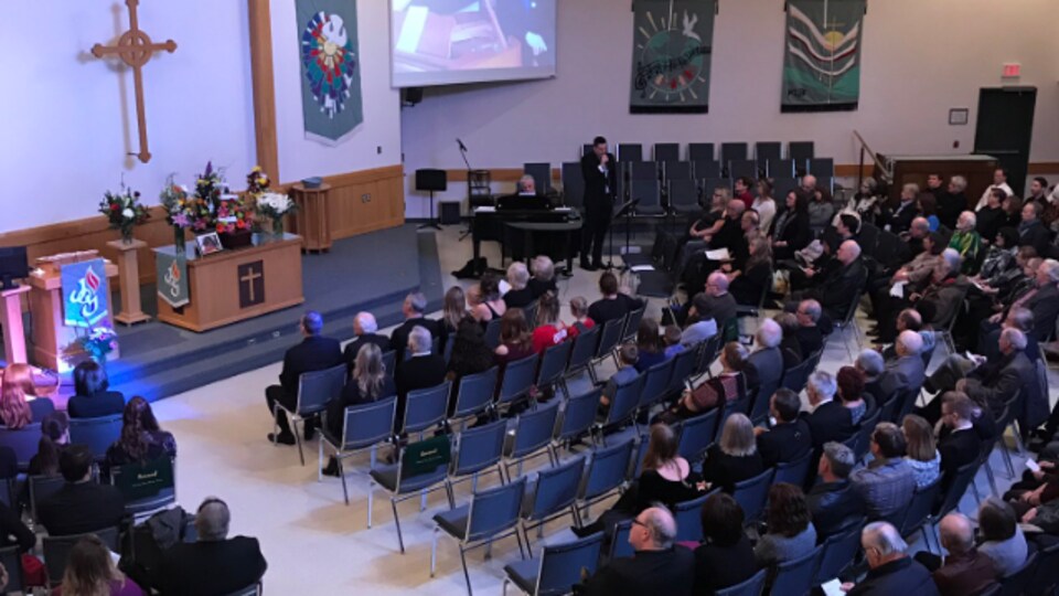 Vue élevée sur les sièges et l'autel d'une église de Saskatoon avec des gens qui sont assis et écoutent un pianiste et un chanteur.