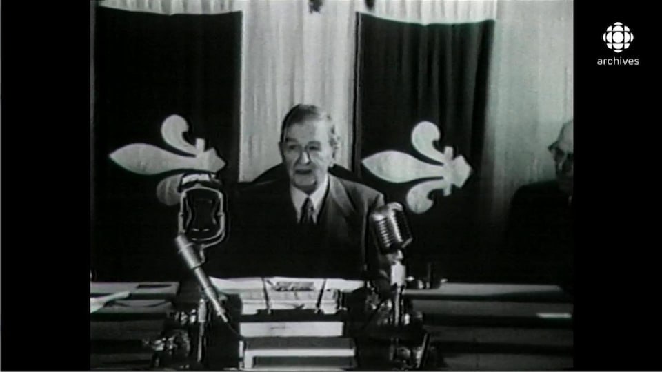 Image en noir et plan de Maurice Duplessis, assis à un bureau massif équipé de micros, avec en arrière-plan un grand drapeau du Québec