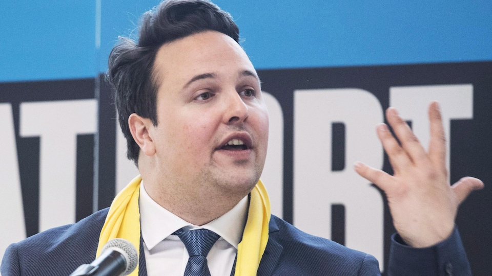 M. Laporte derrière un micro, portant une écharpe jaune.