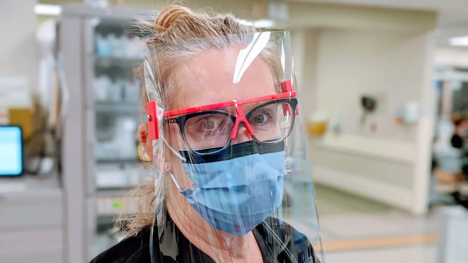 Une femme portant un masque et des lunettes surmontées a le visage couvert d’une pellicule plastique retenue par une forme de lunettes.