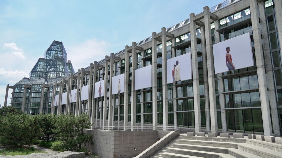 La façade du MBAC sur laquelle on voit un accrochage d'une série de photos sur fond blanc représentant un femme et un homme.