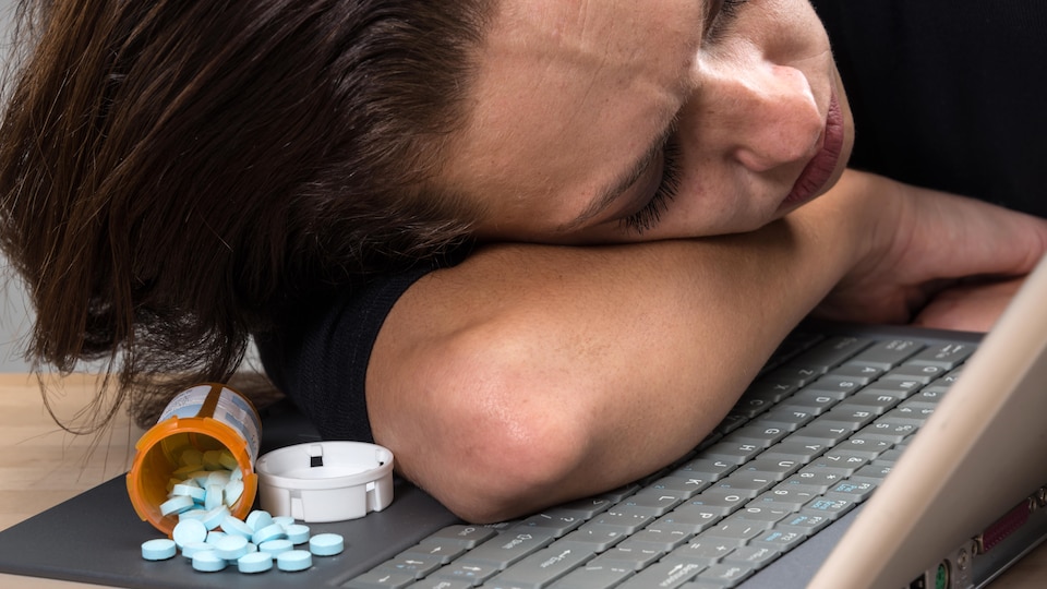 Une femme a la tête couchée sur son ordinateur derrière un pot de pilules.