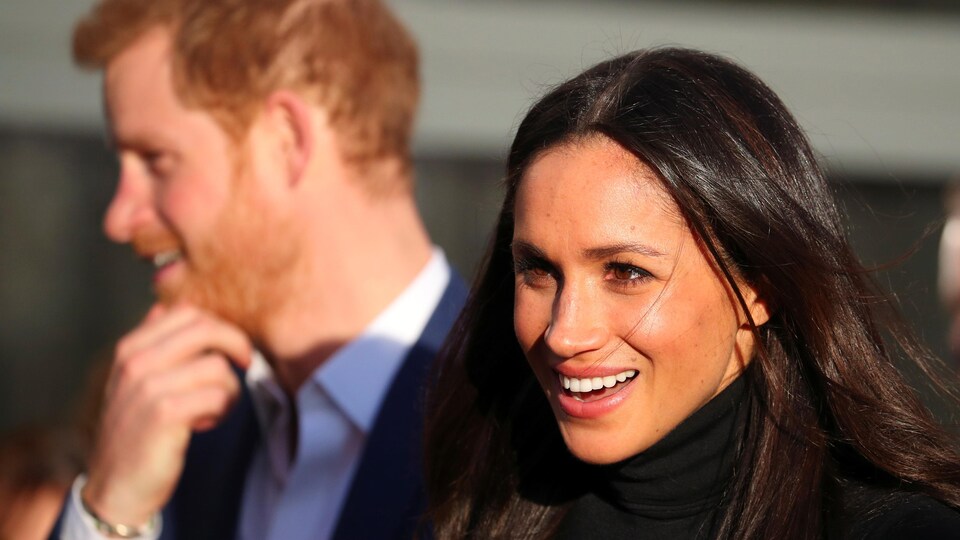 Plan serré de Meghan Markle souriante avec le prince Harry en flou en arrière-plan.