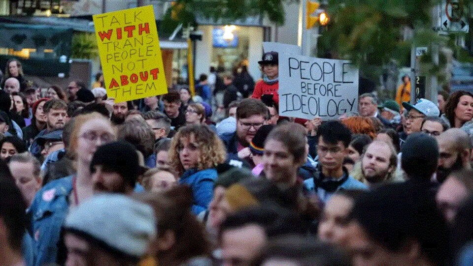 Des manifestants tiennent des affiches avec des messages comme « parle avec les personnes trans, pas à leur sujet ».