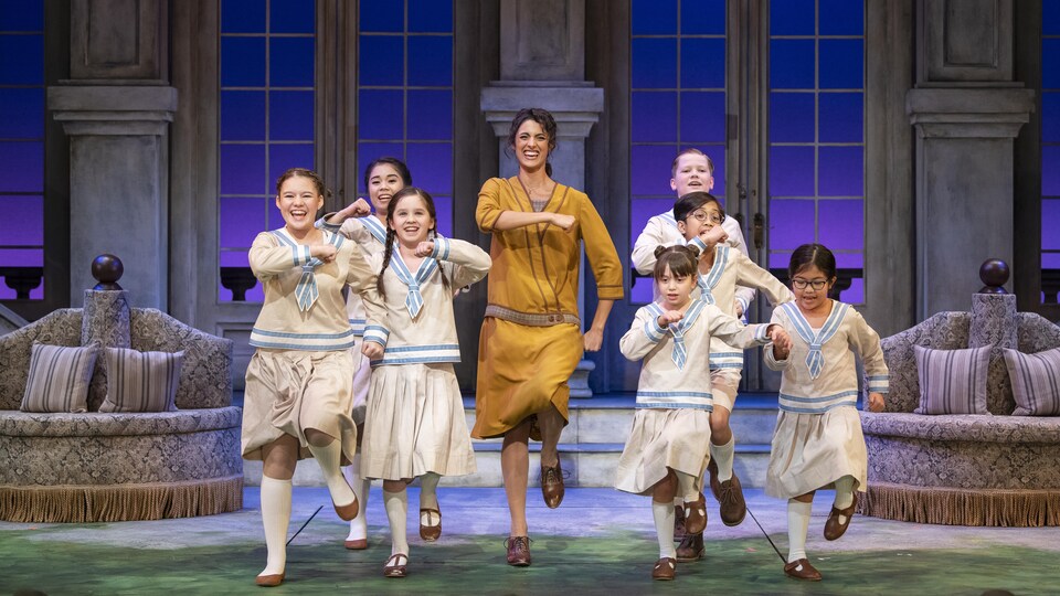 Une femme adulte et sept enfants dansent et chantent dans une scène de la pièce musicale « The Sound of Music ».