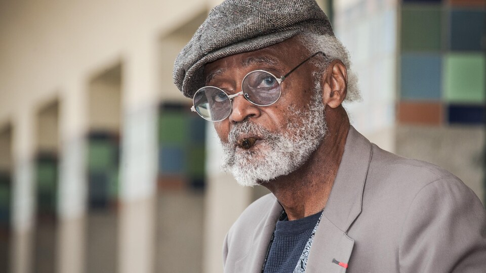 Le réalisateur, d'un âge avancé, porte une barbe blanche, une casquette et des lunettes rondes. Il fume un cigare.