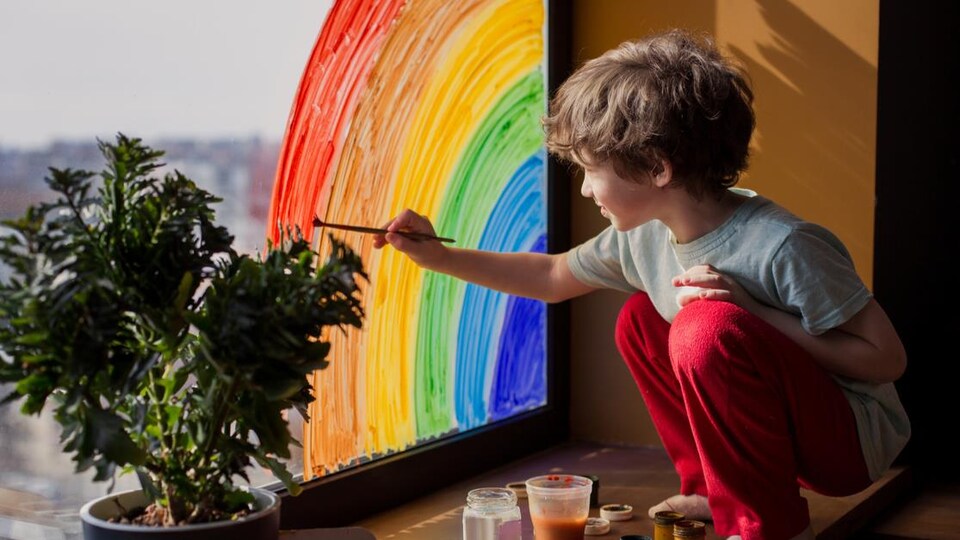 une petit garçon peint un arc-en-ciel sur une vitre.