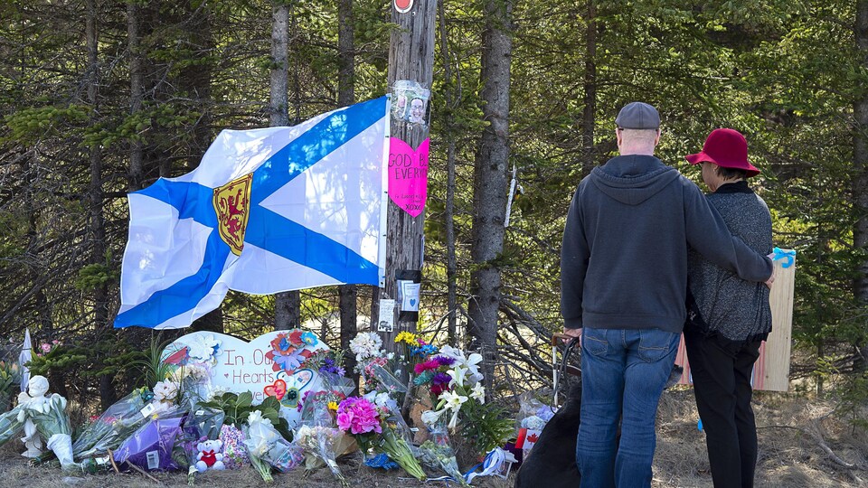 Deux personnes de dos devant un drapeau qui flotte au-dessus de fleurs et de divers objets posés sur le sol.