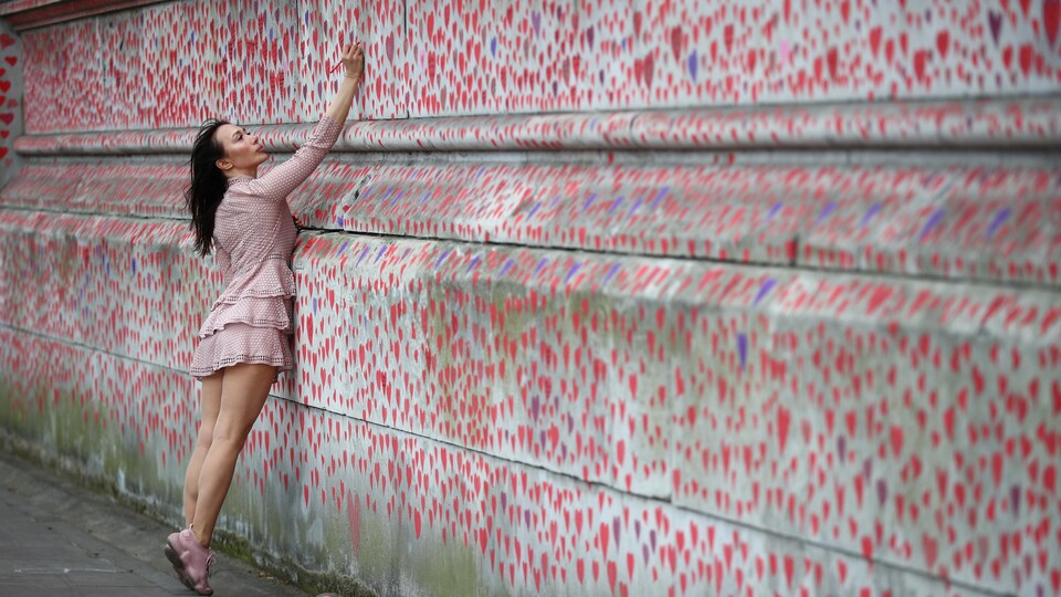 Une femme sur la pointe des pieds écrit un message sur un mur tapissé de coeurs rouges. 
