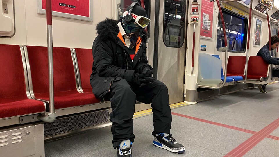 Un homme portant des lunettes de ski, une cagoule et des gants est assis dans un wagon de métro.