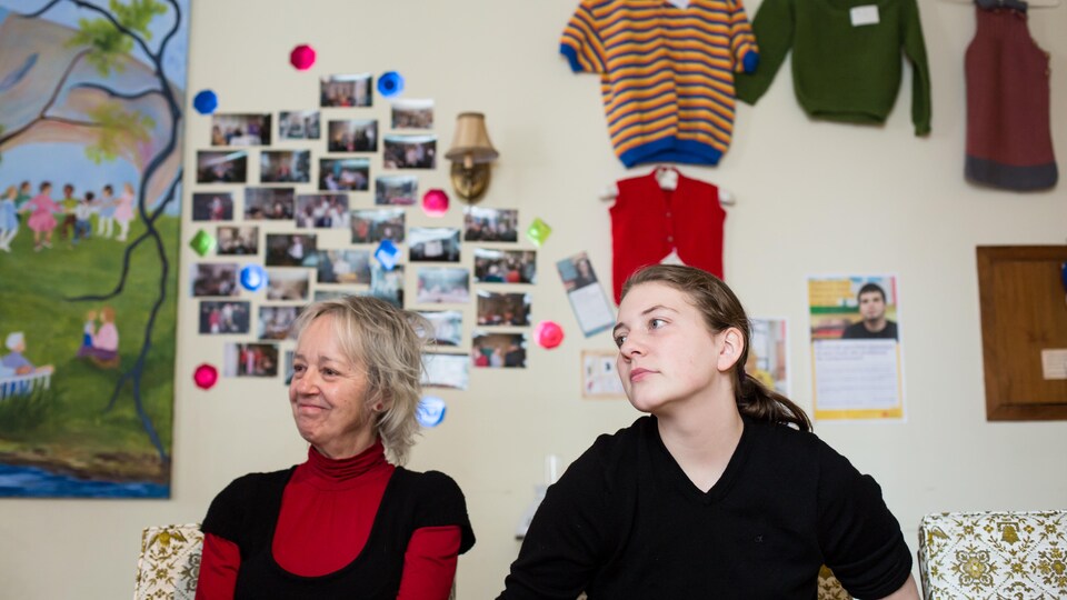 L'aînée Suzanne Lavallée et l'adolescente Lydia De Melo sont assises côte-à-côte devant un mur orné de photos et de vêtements tricotés à la main.