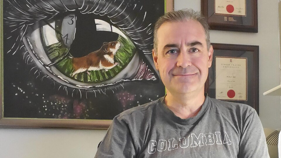 Michael Antle, assis dans son bureau devant un tableau proposant une combinaison d'un oeil, d'un hamster sur une roue et d'une horloge.
