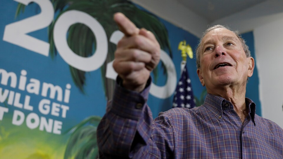 Michael Bloomberg se trouve devant une affiche promotionnelle de sa campagne où on peut lire Miami.