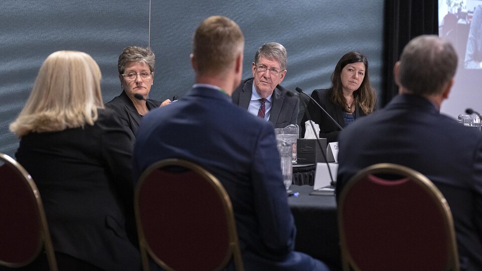 Les trois commissaires assis côte à côte à une table ronde, avec d'autres personnes vues de dos, assises face à eux.