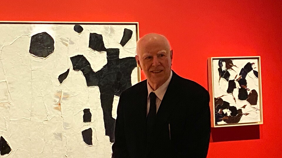 L'homme porte un veston et une cravate. Il est devant deux tableaux et il sourit. 