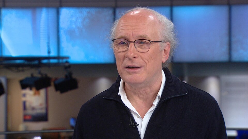 Un homme portant des lunettes est sur un plateau de télévision. Il sourit légèrement.