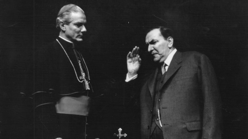 Un cardinal et un homme en veston discutent sur une scène de théâtre.