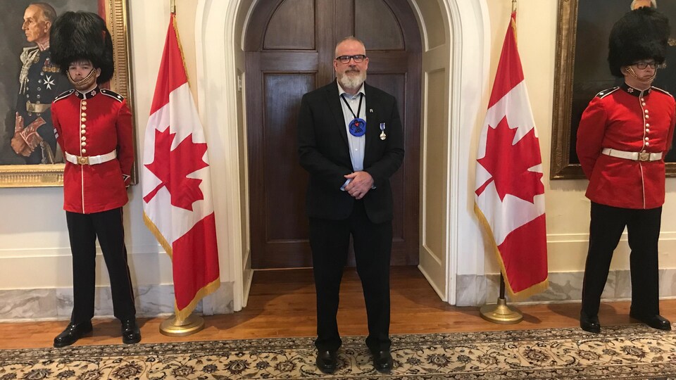 Michel Bourbonnière à Rideau Hall portant sa médaille debout entre deux drapeaux canadiens et deux membres de la Gendarmerie royale du Canada.