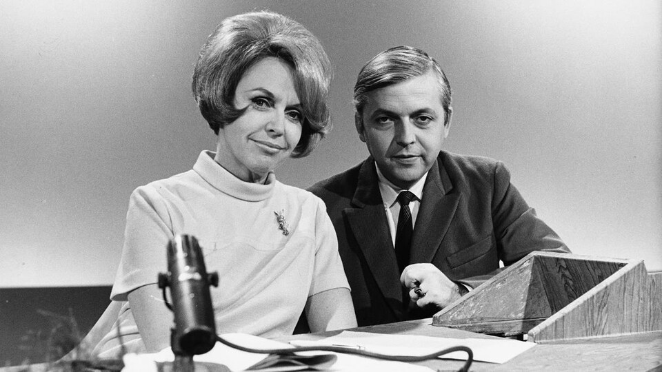 Dans un studio de télévision, les animateurs Michelle Tisseyre et Wilfrid Lemoine sont assis à un pupitre surmonté d'un micro de table.