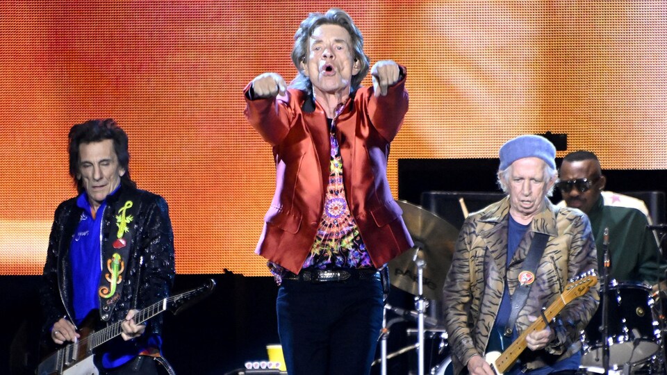 Mick Jagger pointe la foule sur scène, entouré de deux guitaristes et d'un batteur en arrière-plan. 
