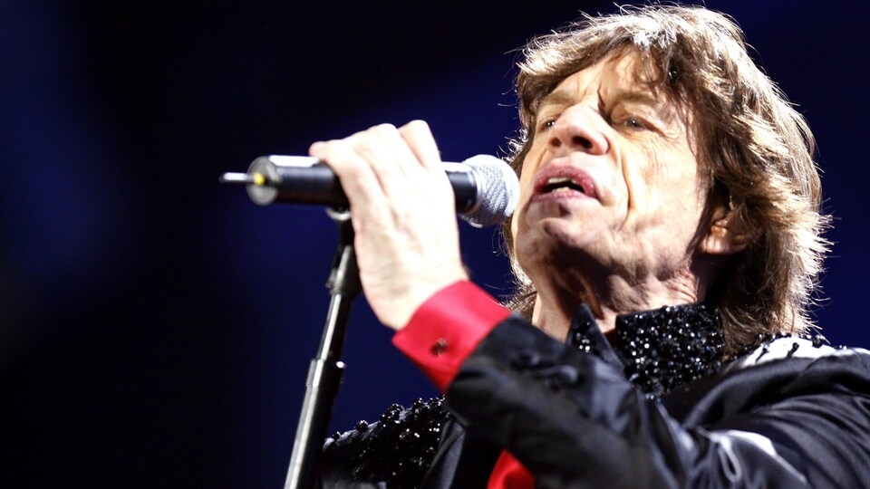 Mick Jagger, sur scène, tient un micro et chante.