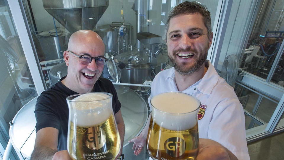 Le fondateur de la Microbrasserie Charlevoix, Frédérick Tremblay, et le brasseur Nicolas Marrant lèvent des verres de bière contenant La G7, devant des cuves de brassage dans la microbrasserie.