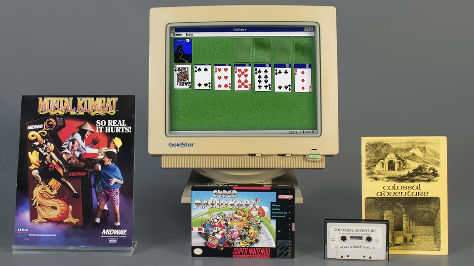 Un photo montrant une affiche pour la borne d'arcade de Mortal Kombat, la pochette américaine du jeu Super Mario Kart, une cassette audio et un livret jaune de Colossal Cave Adventure et un vieil écran d'ordinateur affichant une vieille version du jeu Microsoft Solitaire.