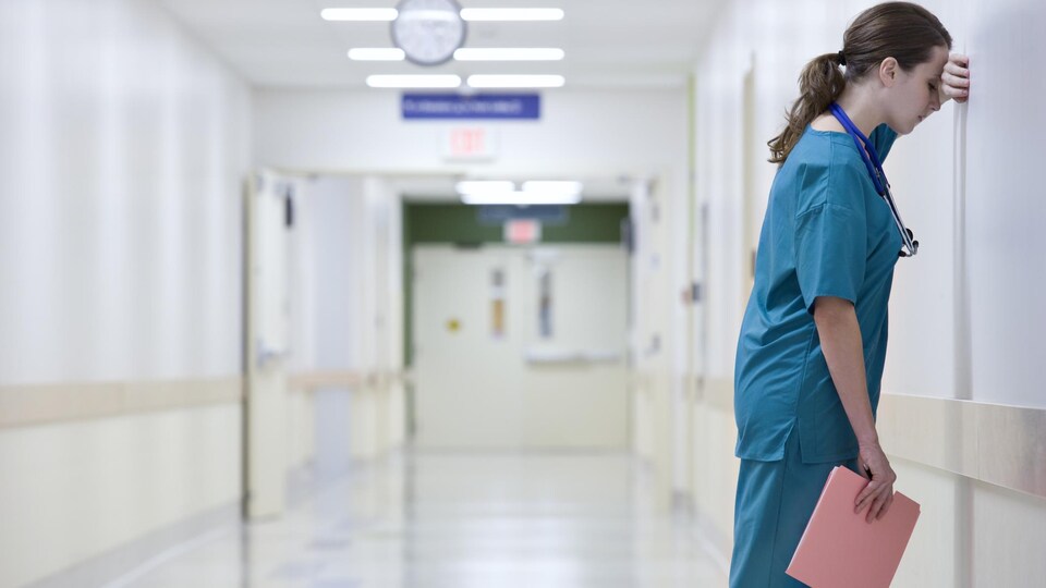 Une professionnelle de la santé est appuyée contre un mur dans un couloir d'hôpital.