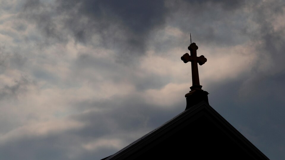 Des nuages noires se glissent à l'arrière-plan d'une croix sur le toit d'une église catholique.
