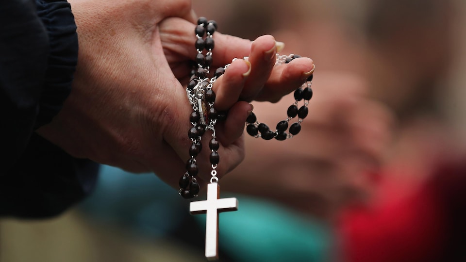 Les mains jointes, une paroissienne tient un chapelet avec, au bout, une croix bien visible au bout de la chaîne.