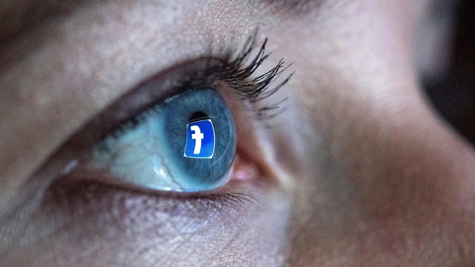 Une femme se tient devant un écran d'ordinateur alors que le logo du réseau social Facebook se réflète dans son œil. 