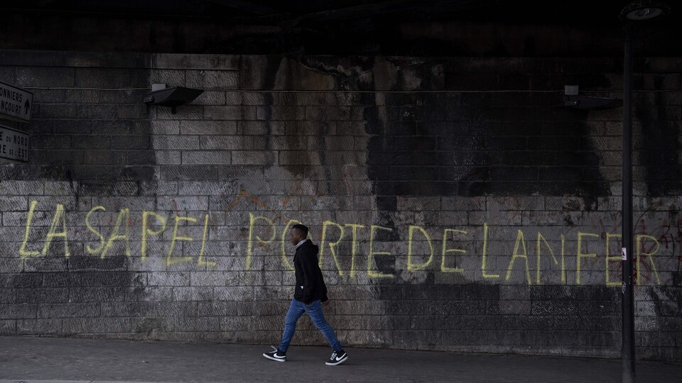Un jeune homme marche près d'un mur où il est écrit : « La sapel porte de lanfer ».