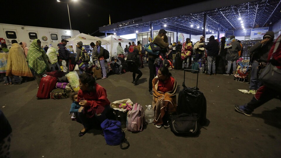 Des adultes s'enveloppant de couvertures et quelques enfants attendent avec leurs bagages à l'extérieur en soirée le feu vert des autorités pour entrer en Équateur