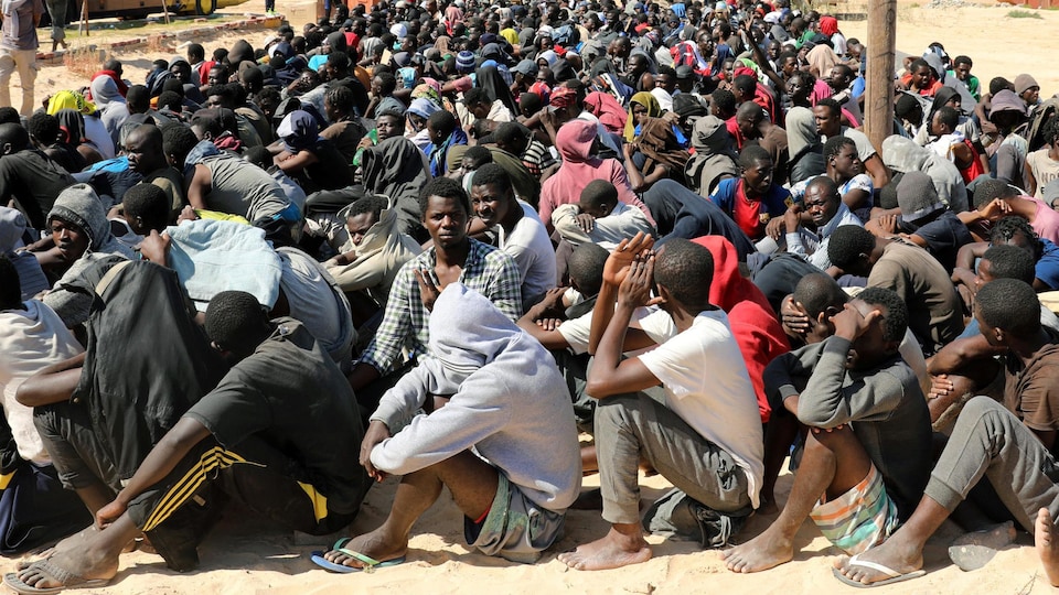 Des centaines d'hommes sont assis parterre dans le sable en rang serré.