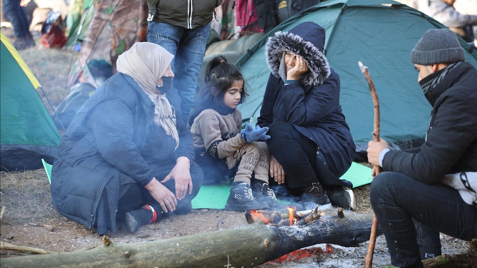 Des migrants sont assis dans un camp, près d'un feu.