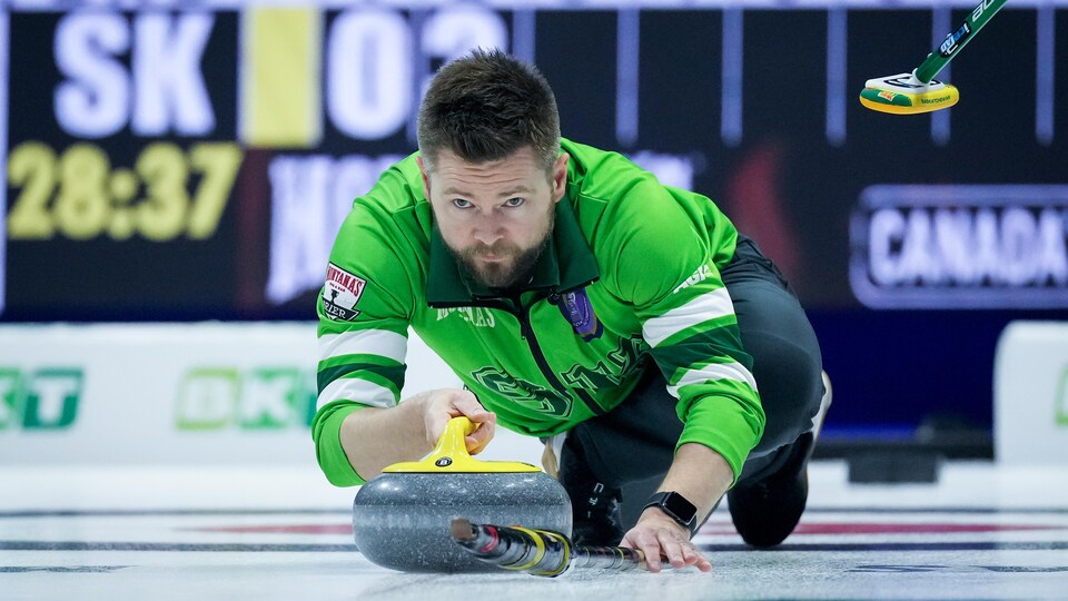 Un joueur de curling vêtu de vert envoie une pierre en s'appuyant sur son bâton.