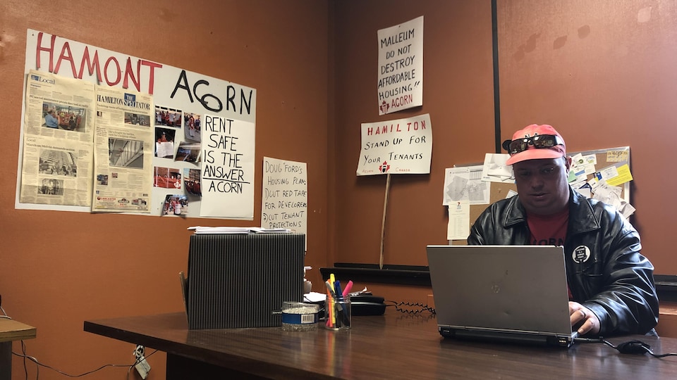 Un homme travaille à un bureau. On voit derrière lui des pancartes portant des messages à propos des logements sociaux de Hamilton.