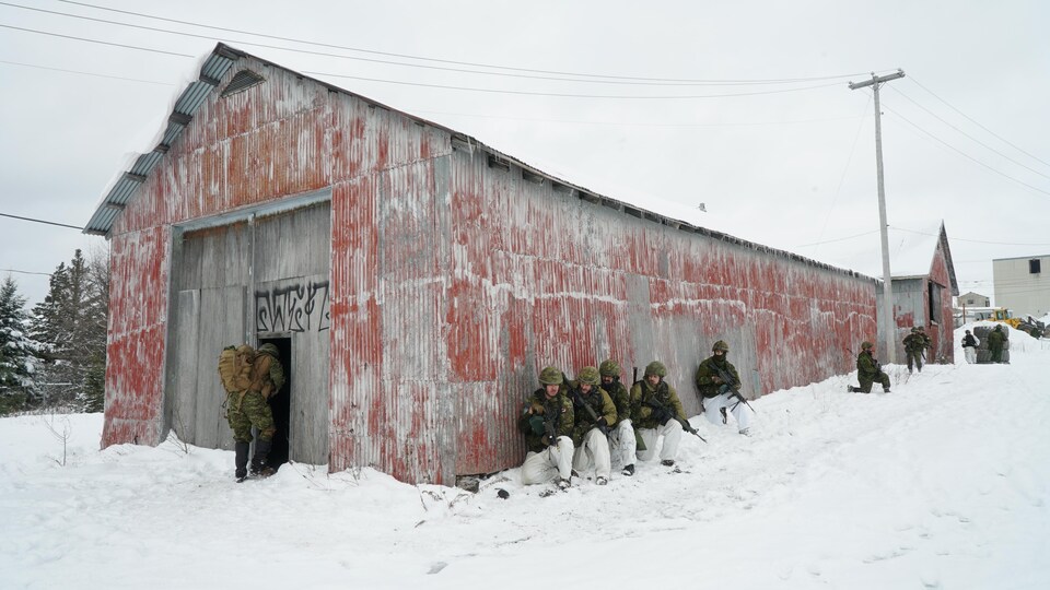 Des militaires longent le mur de tôle d'un vieux bâtiment. Ils sont immobiles. Un autre militaire tout près entre dans le bâtiment. 